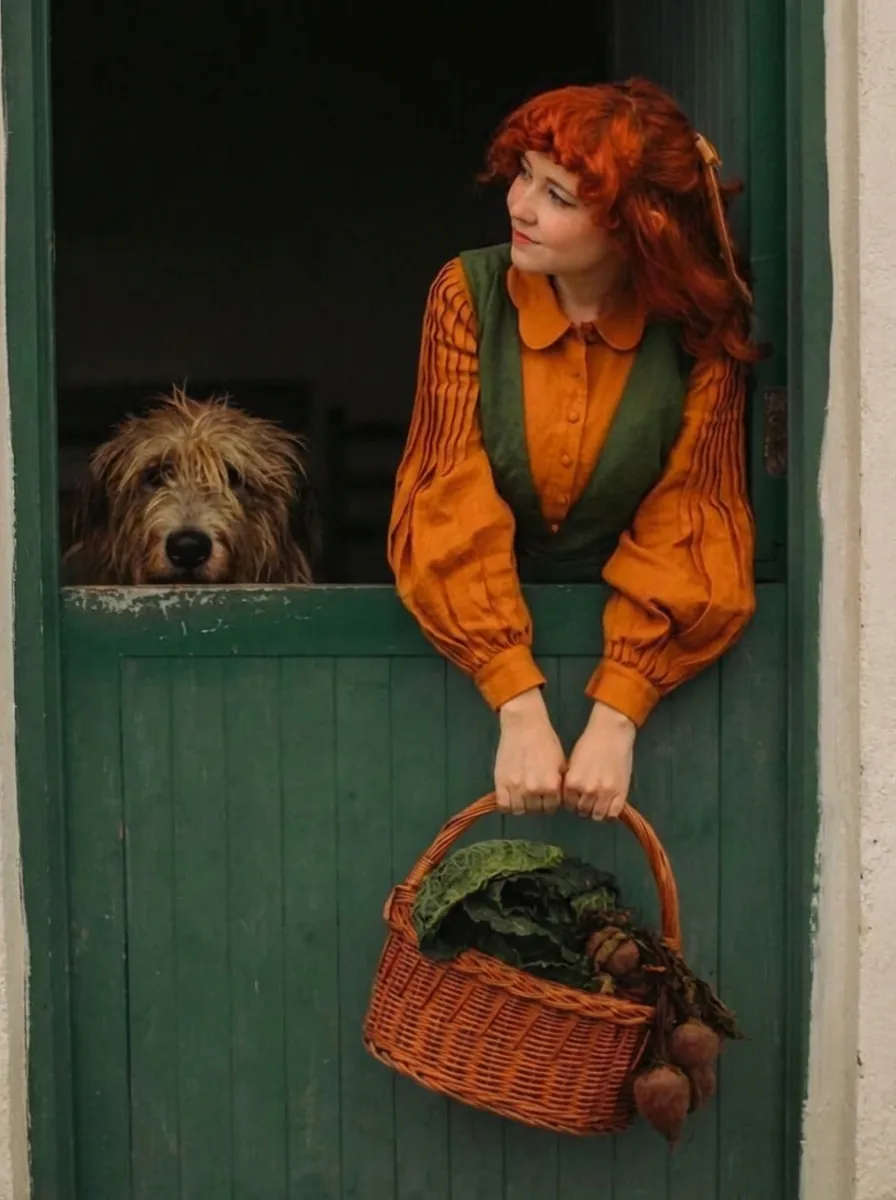 A woman with vibrant red hair and a shaggy dog lean over a dark green stable door. She wears a mustard-orange blouse and a green vest while holding a wicker basket filled with beets and kale, exemplifying the rich, earthy, and warm colors of the Autumn season.