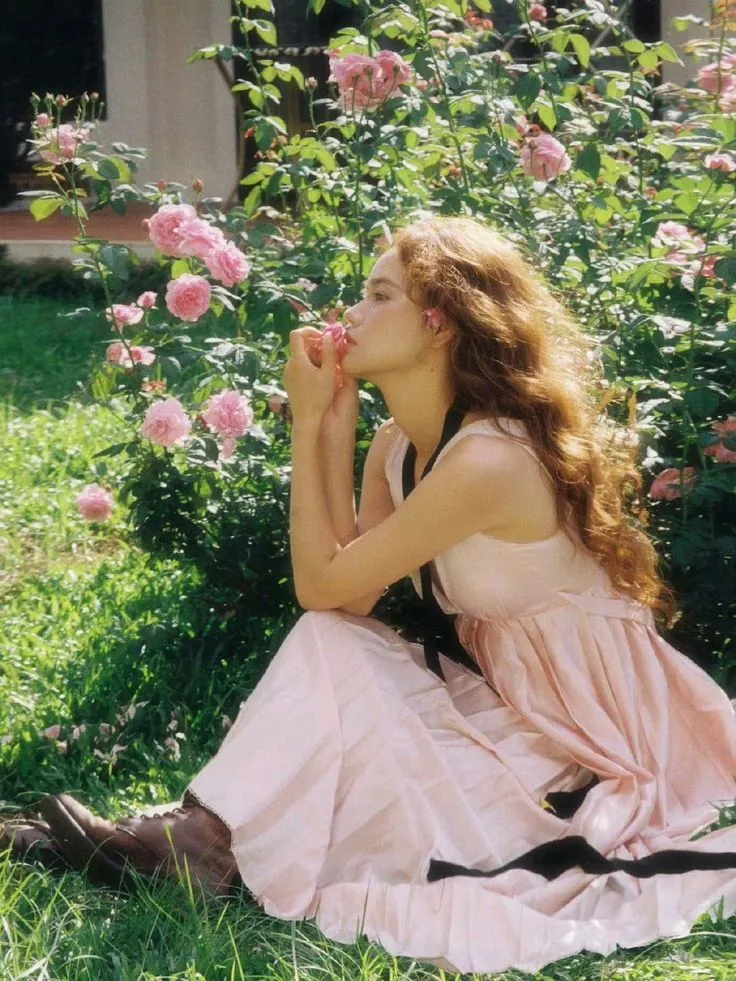 A young woman with long, warm strawberry-blonde hair sits in a sunlit garden surrounded by pink rose bushes. She is wearing a soft pale pink dress and smelling a rose, representing the fresh, warm, and golden aesthetic of the Spring color season.